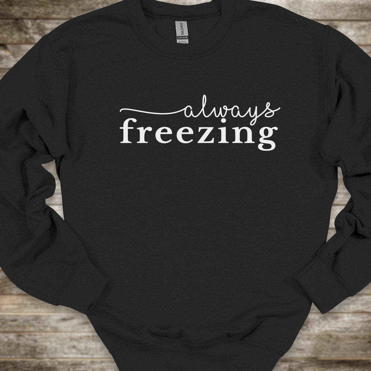 A black crewneck sweatshirt with the phrase 'always freezing' printed in white across the chest, laid flat on a rustic wooden background.
