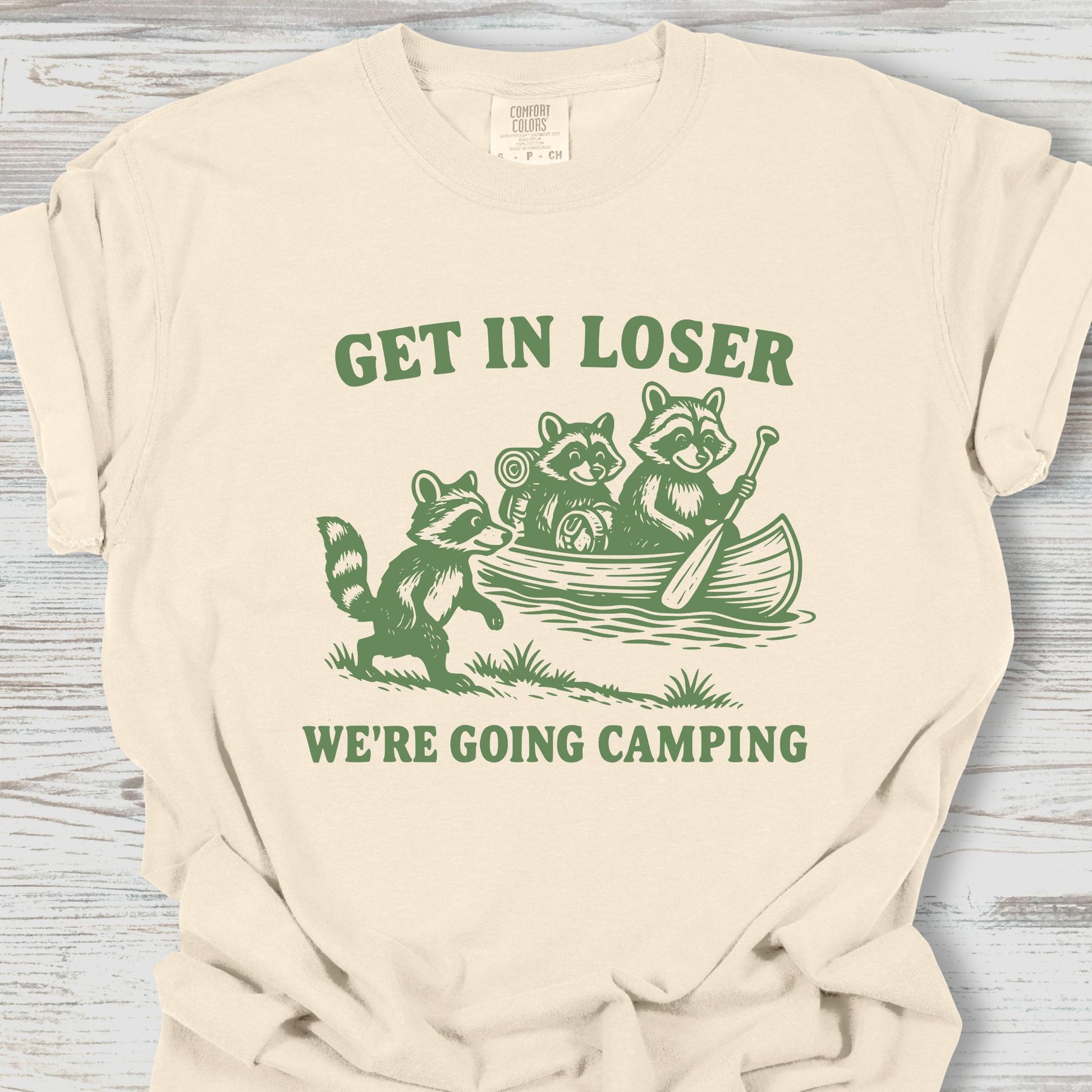 Beige t-shirt with green graphic and text 'Get in loser, we're going camping' on a wooden surface