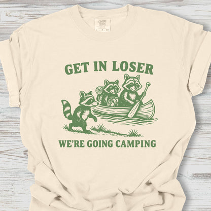 Beige t-shirt with green graphic and text 'Get in loser, we're going camping' on a wooden surface