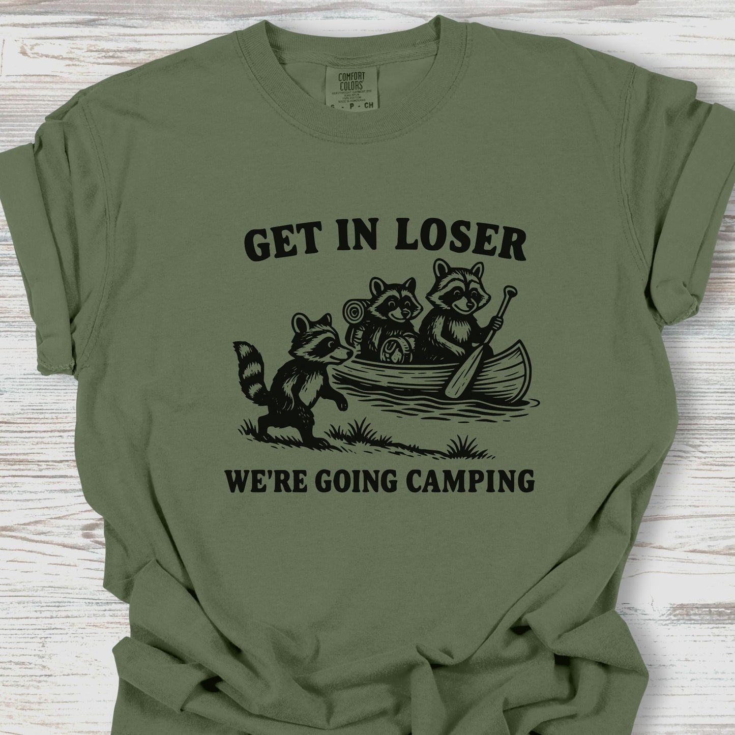 Green t-shirt with raccoon graphic and text 'Get in loser, we're going camping' on a wooden surface.