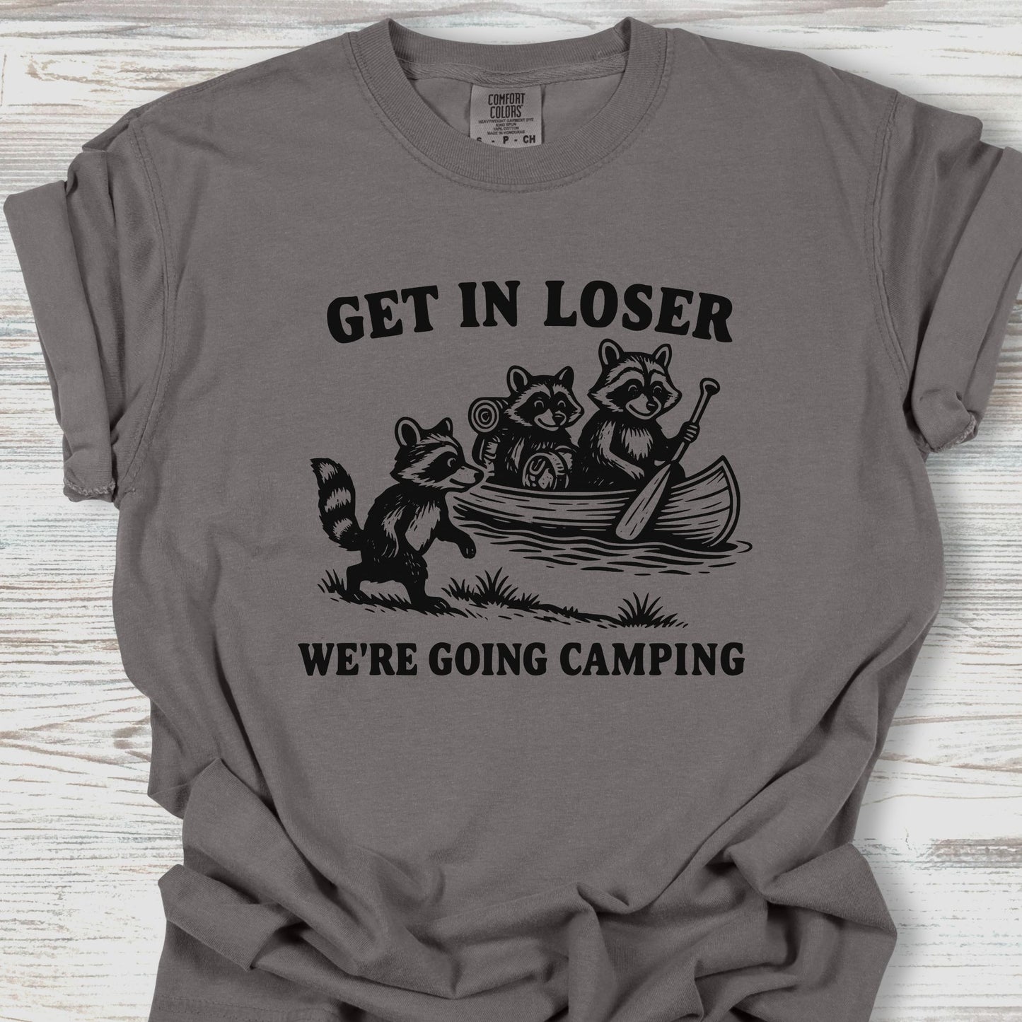 Gray t-shirt with raccoon graphic and text 'Get in loser, we're going camping' on a wooden surface.