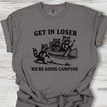 Gray t-shirt with raccoon graphic and text 'Get in loser, we're going camping' on a wooden surface.