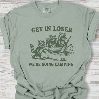 Sage green t-shirt with raccoon graphic and text 'Get in loser, we're going camping' on a wooden surface.