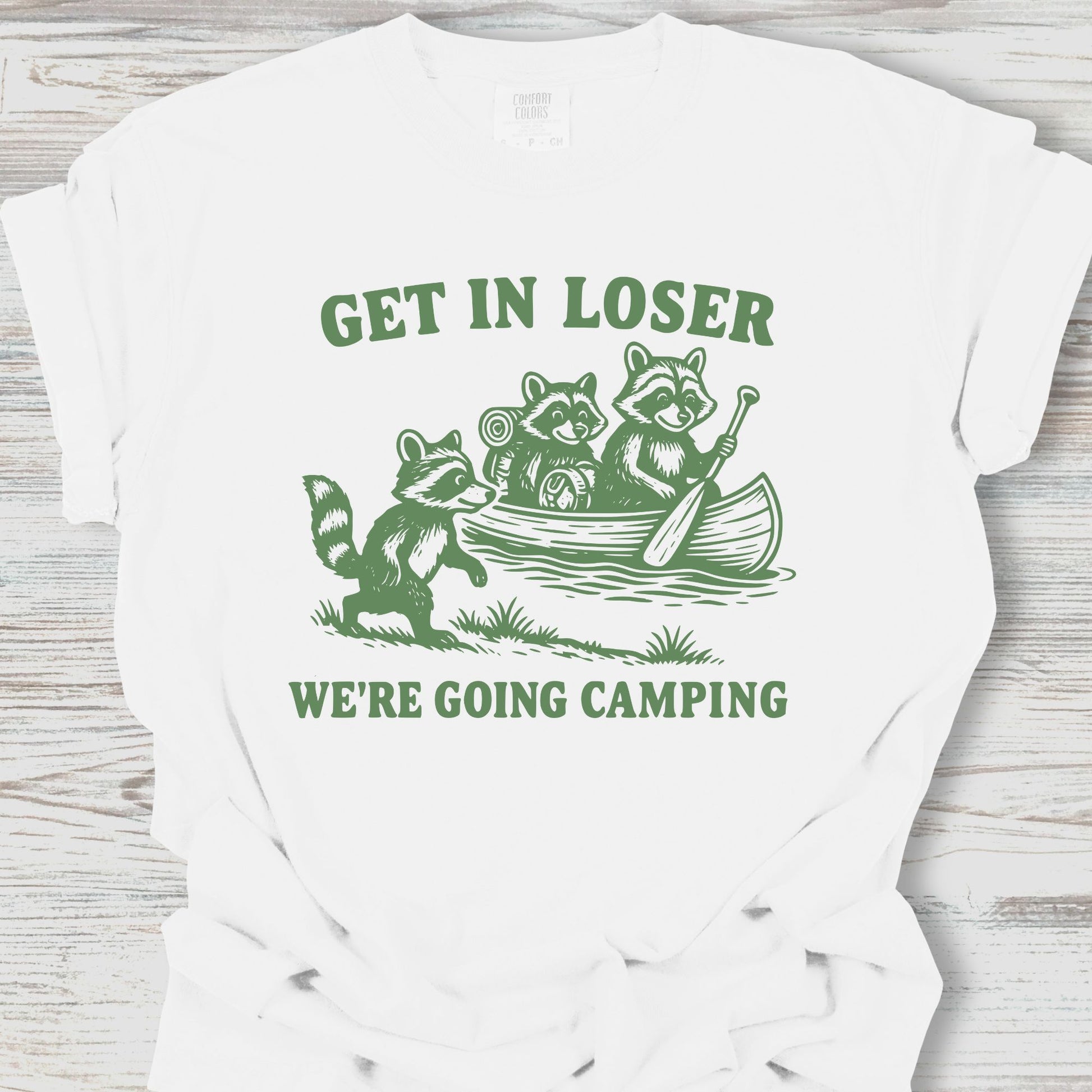 White t-shirt with green graphic and text 'Get in Loser, We're Going Camping' on a wooden surface.