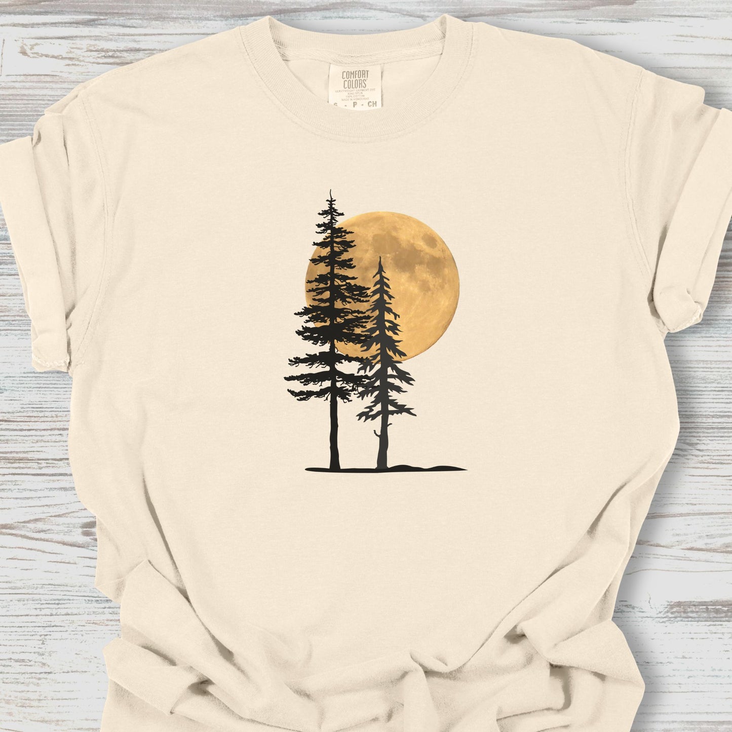 Beige t-shirt with a graphic of three trees and a moon on a wooden surface