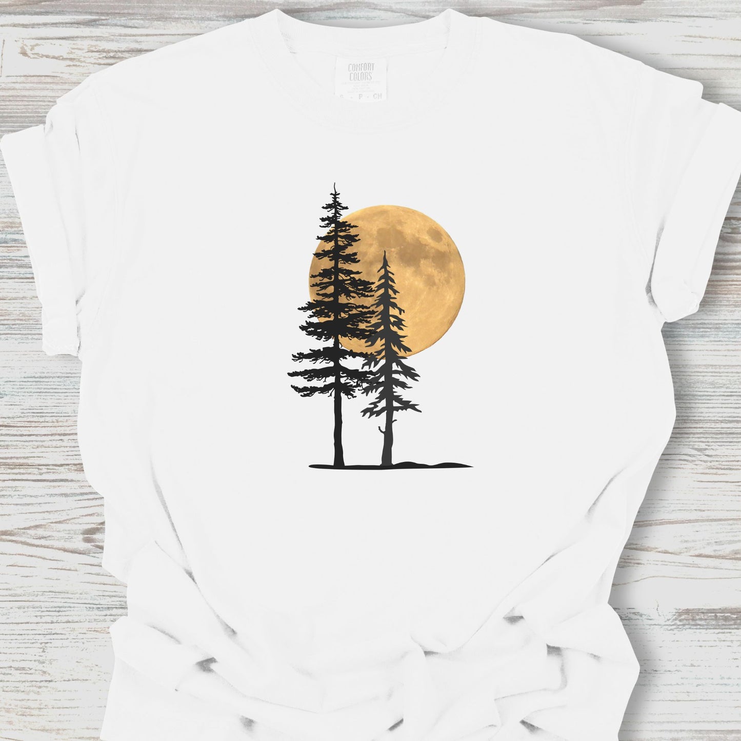 White t-shirt with a graphic of two trees and a full moon on a wooden surface