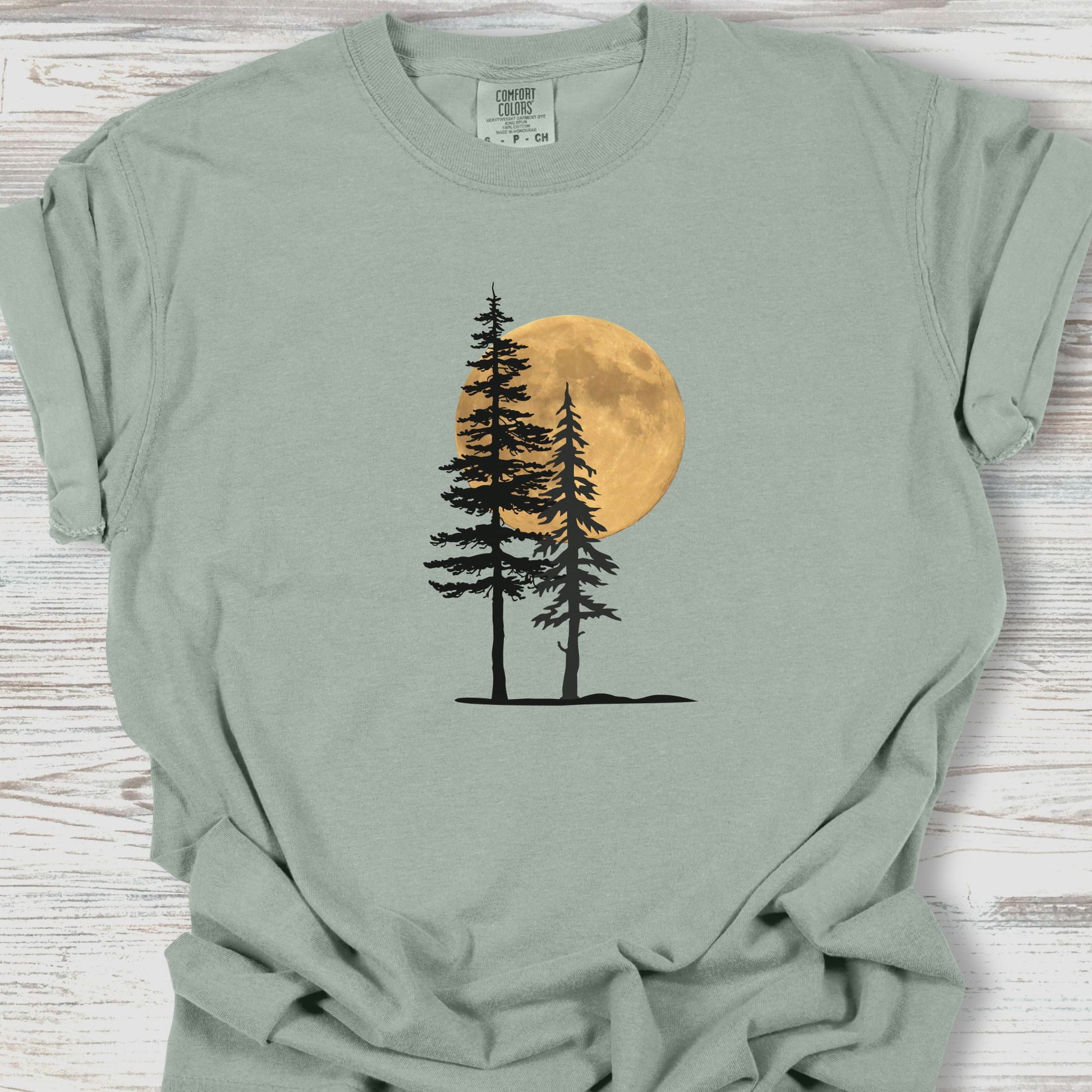 Sage green t-shirt with a graphic of trees and a moon on a wooden surface