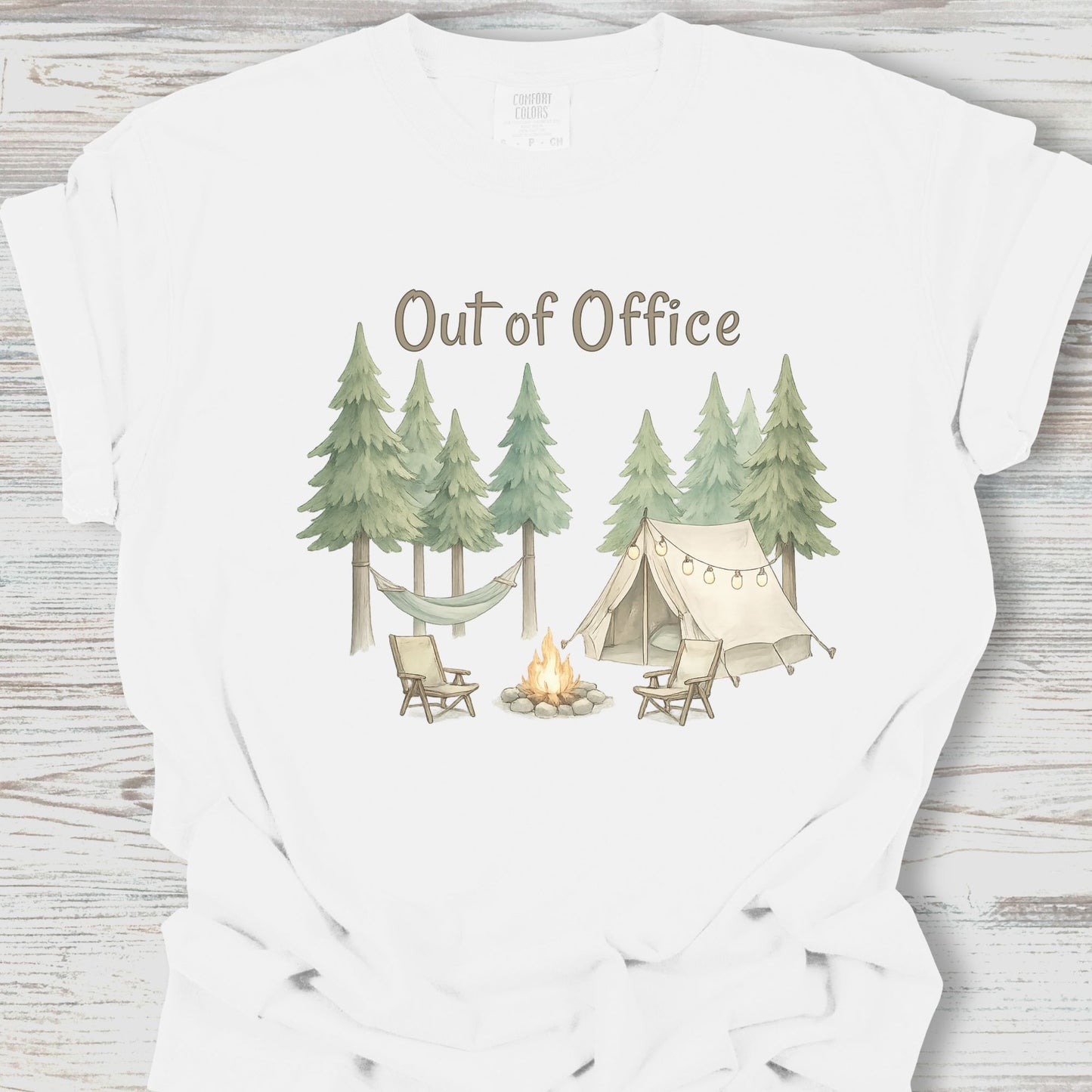 White t-shirt with a camping scene graphic and 'Out of Office' text on a wooden surface.