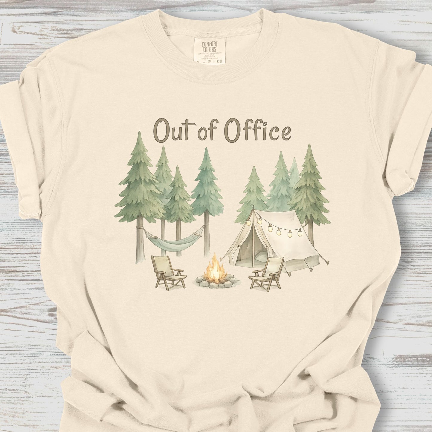 Beige t-shirt with a camping graphic and 'Out of Office' text on a wooden surface