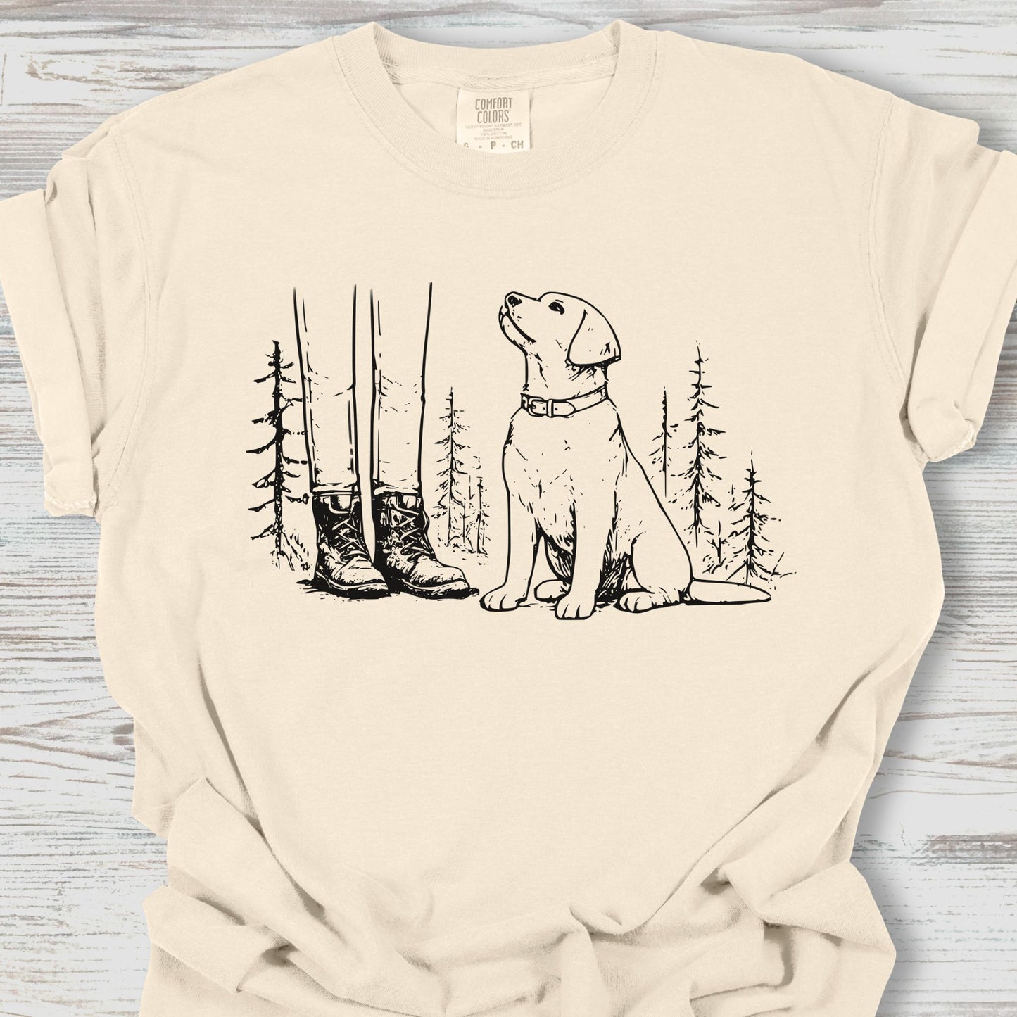 Beige t-shirt with a graphic of a dog and boots in a forest on a wooden surface