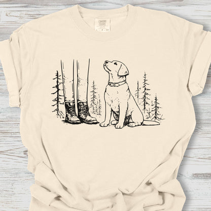 Beige t-shirt with a graphic of a dog and boots in a forest on a wooden surface