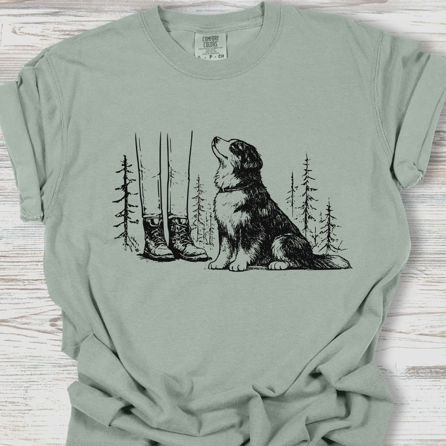 Sage green t-shirt with a graphic of a dog, boots, and trees on a wooden surface