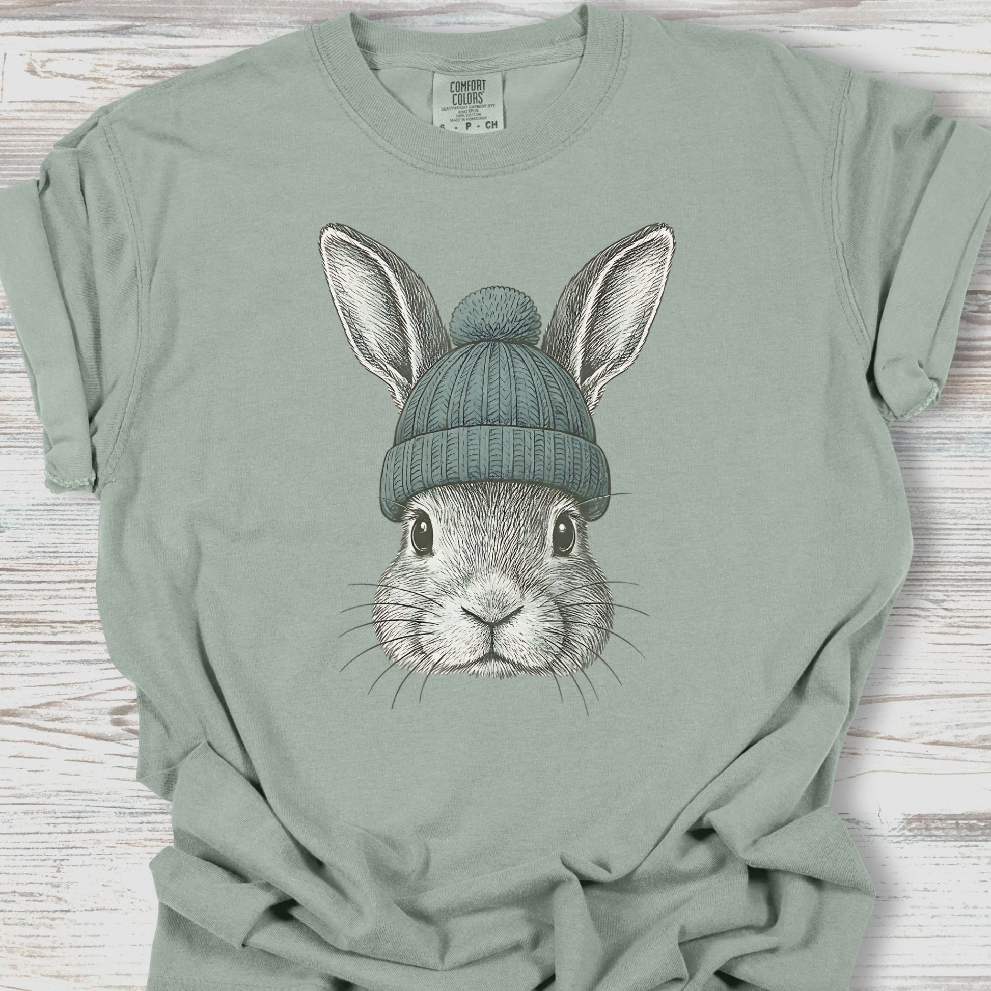 Sage green t-shirt with a graphic of a rabbit wearing a beanie on a wooden surface