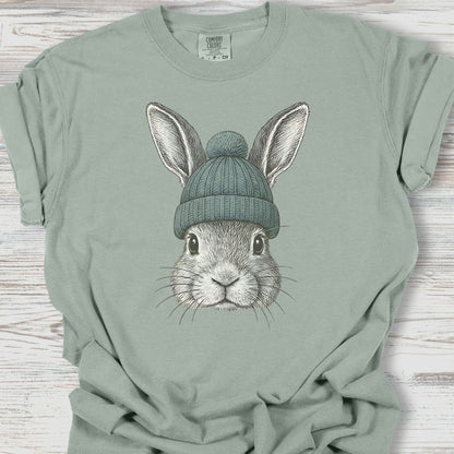 Sage green t-shirt with a graphic of a rabbit wearing a beanie on a wooden surface