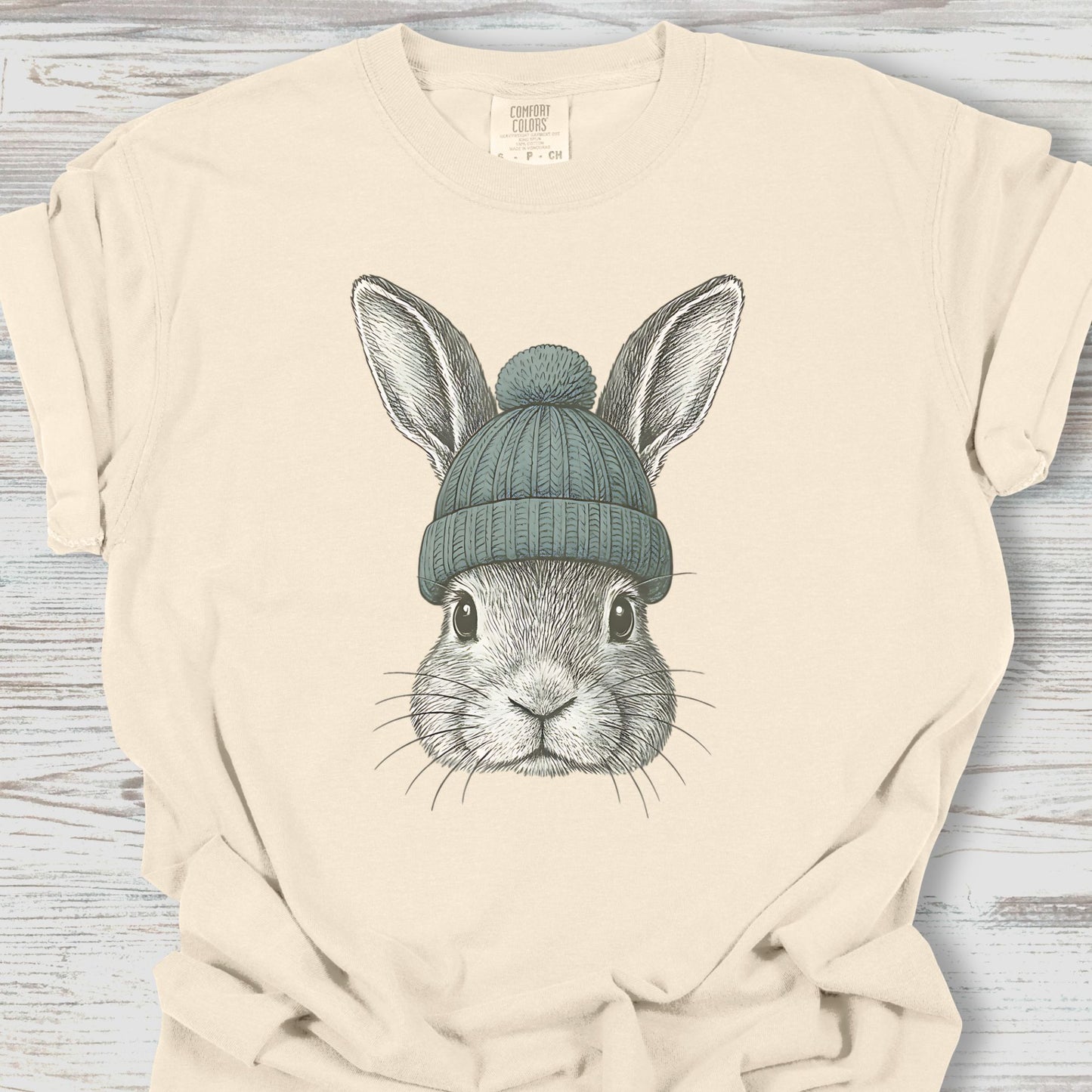 Beige t-shirt with a graphic of a rabbit wearing a green beanie on a wooden surface