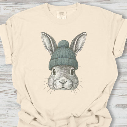 Beige t-shirt with a graphic of a rabbit wearing a green beanie on a wooden surface