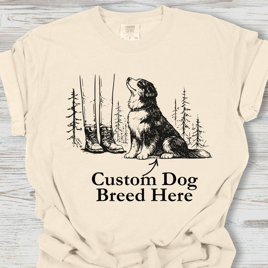 Beige t-shirt with a graphic of a dog, boots, and trees on a wooden surface