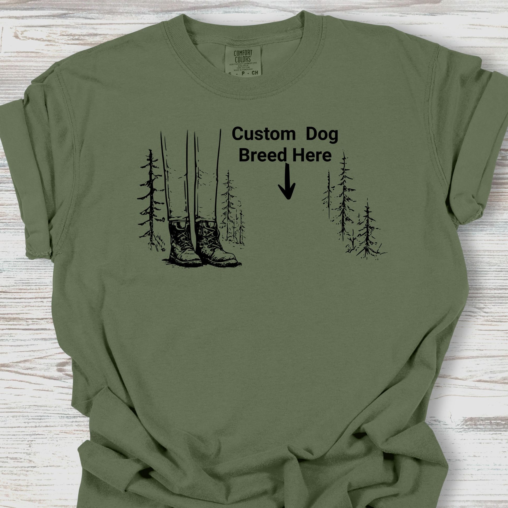 Green t-shirt with graphic of boots and trees and text 'Custom Dog Breed Here' on a wooden surface.