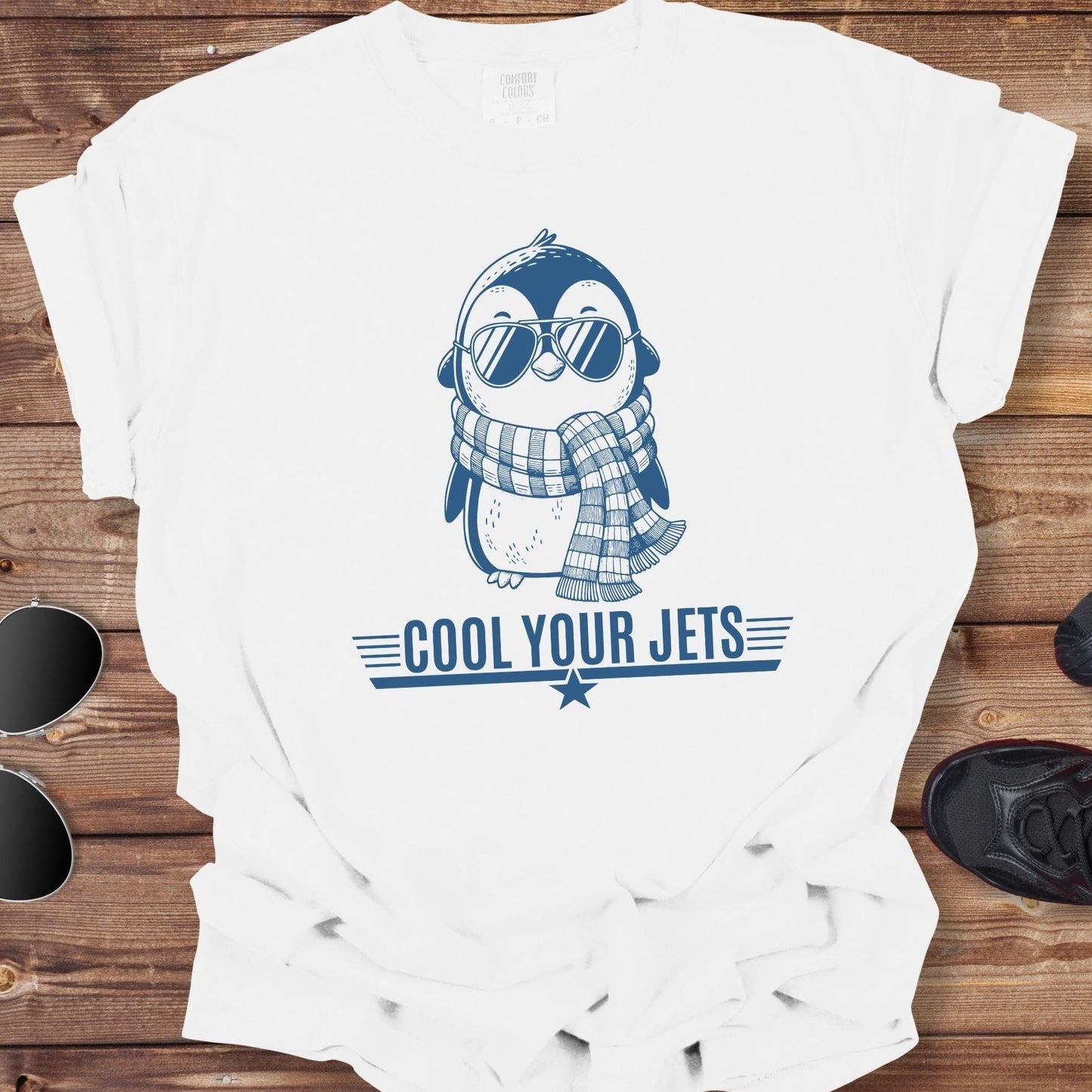 White t-shirt with a penguin graphic and 'Cool Your Jets' text on a wooden surface, accompanied by sunglasses and shoes.
