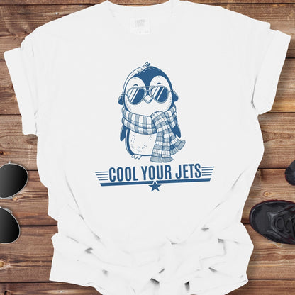 White t-shirt with a penguin graphic and 'Cool Your Jets' text on a wooden surface, accompanied by sunglasses and shoes.