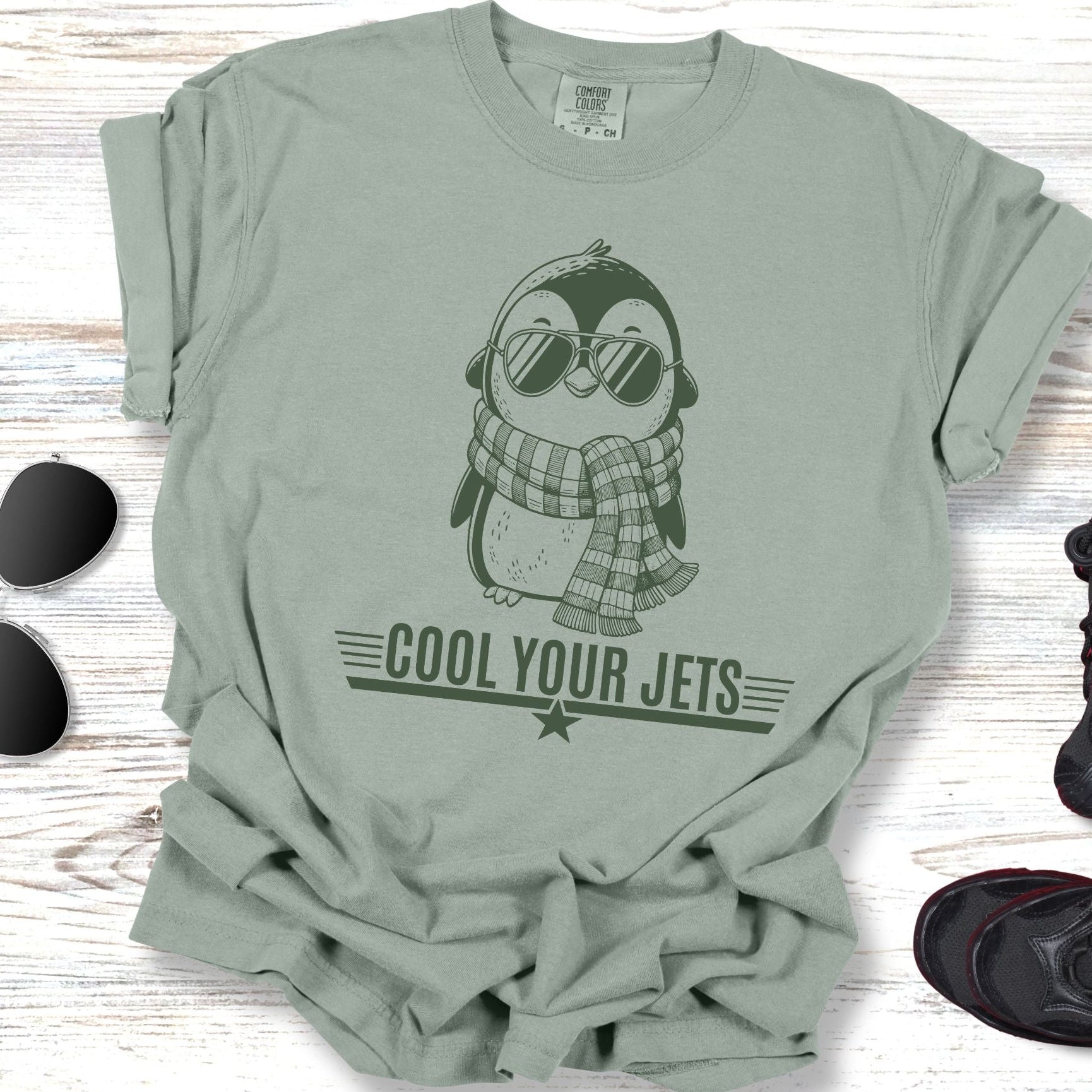 Green t-shirt with penguin graphic and 'Cool Your Jets' text on a wooden surface with sunglasses and shoes.