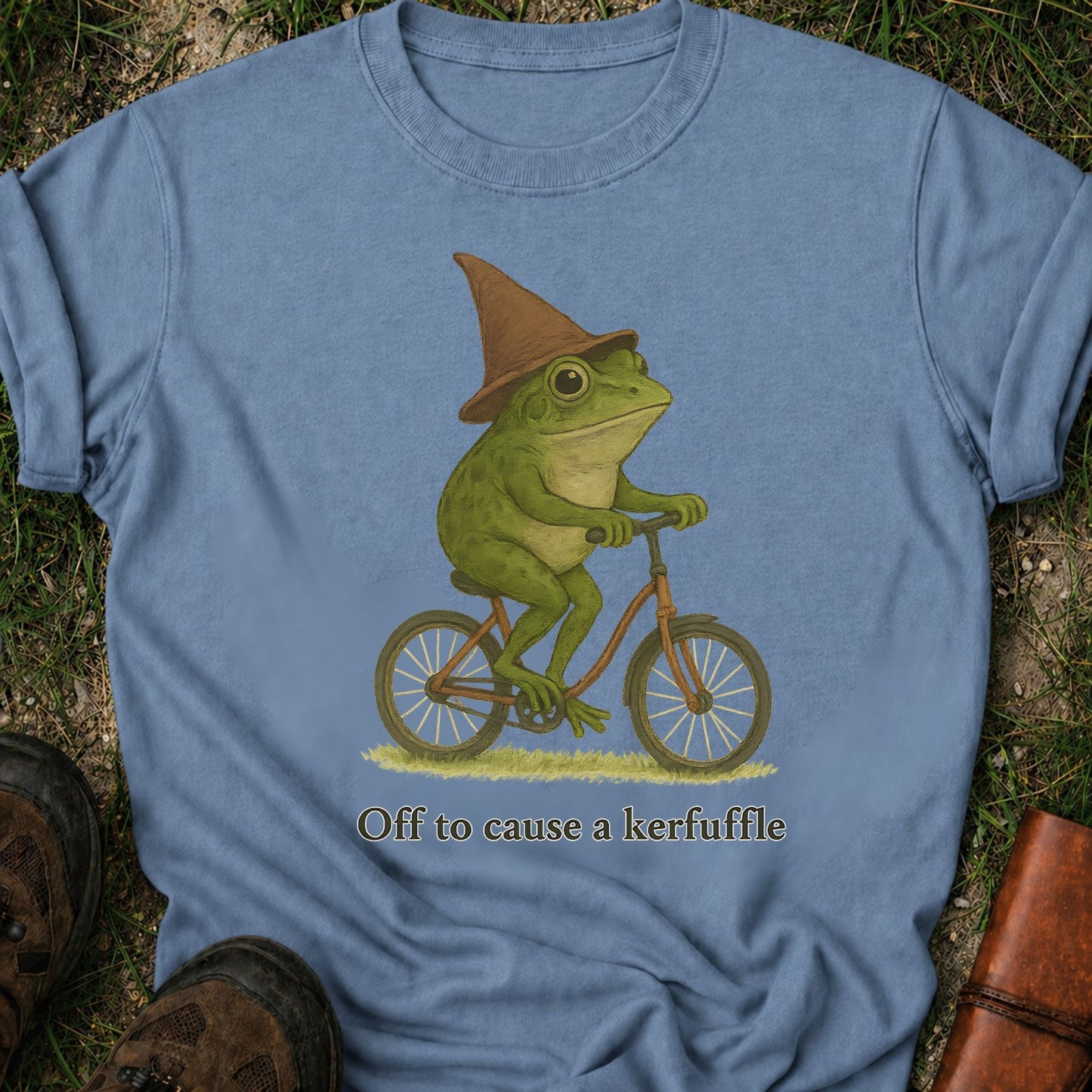 Blue t-shirt with a frog wearing a witch hat riding a bicycle graphic and text 'Off to cause a kerfuffle'.