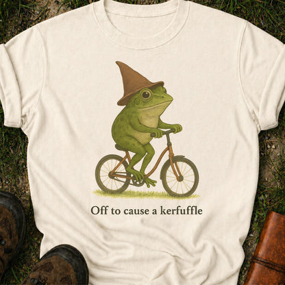 Ivoryt-shirt with a graphic of a frog riding a bike and text 'Off to cause a kerfuffle' on grass.