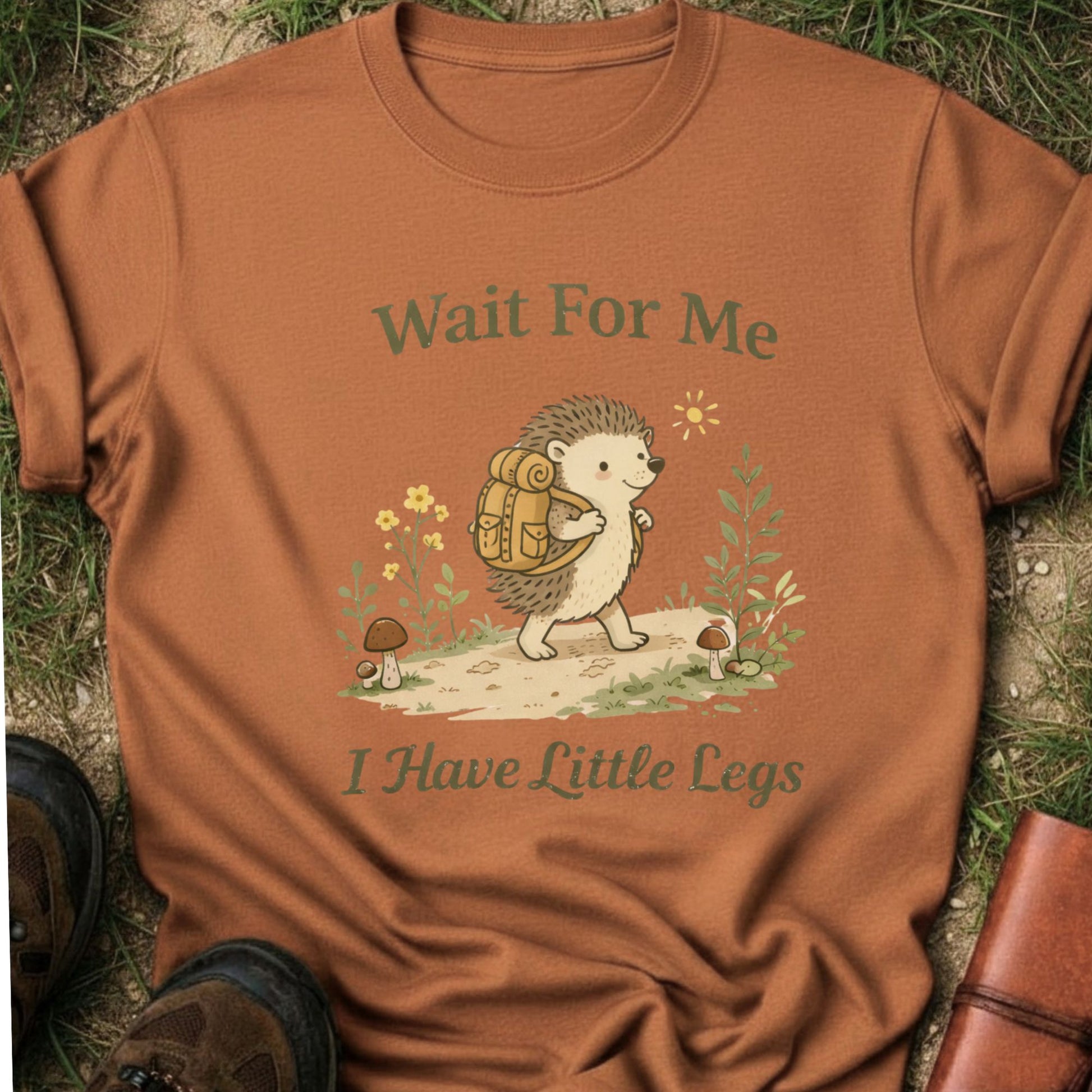 Orange T-shirt with a hedgehog with a hiking backpack strolls through a wildflower meadow, surrounded by mushrooms and greenery. Text reads "Wait For Me, I Have Little Legs."