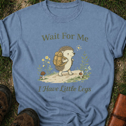 Blue Tshirt with a hedgehog with a hiking backpack strolls through a wildflower meadow, surrounded by mushrooms and greenery. Text reads "Wait For Me, I Have Little Legs."