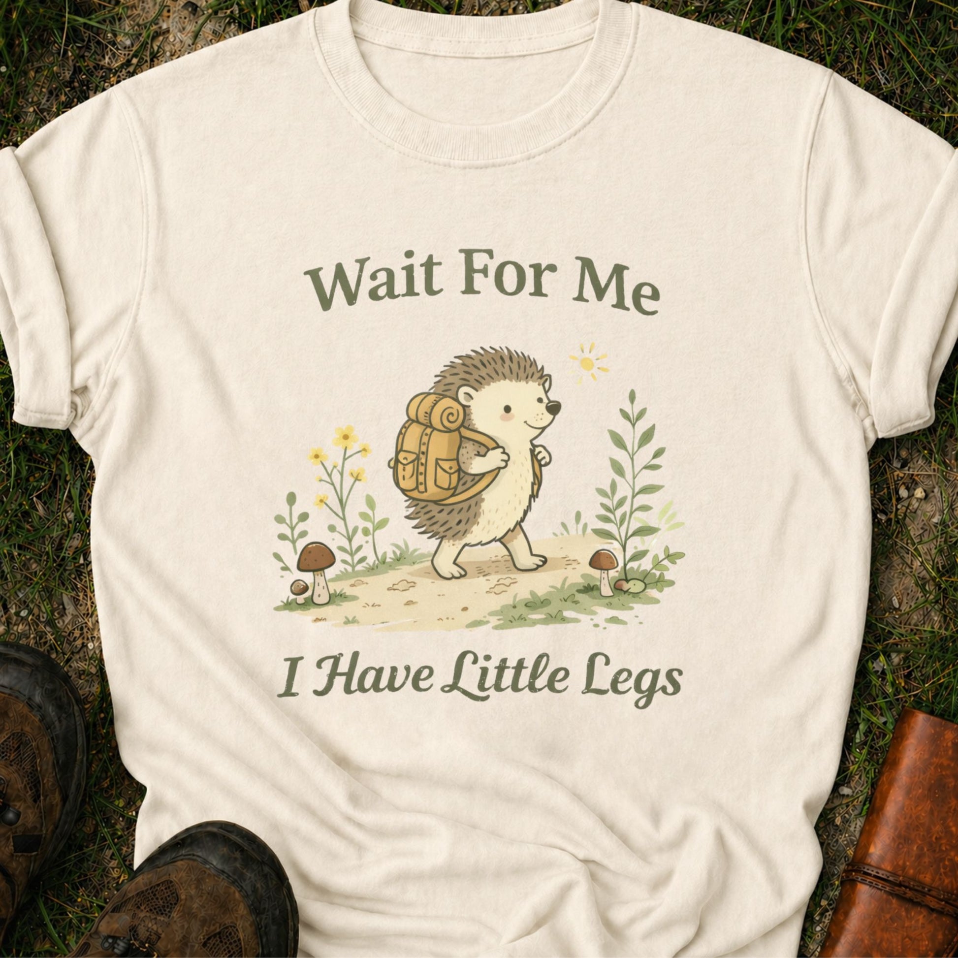Ivory Tshirt with hedgehog with a hiking backpack strolls through a wildflower meadow, surrounded by mushrooms and greenery. Text reads "Wait For Me, I Have Little Legs."