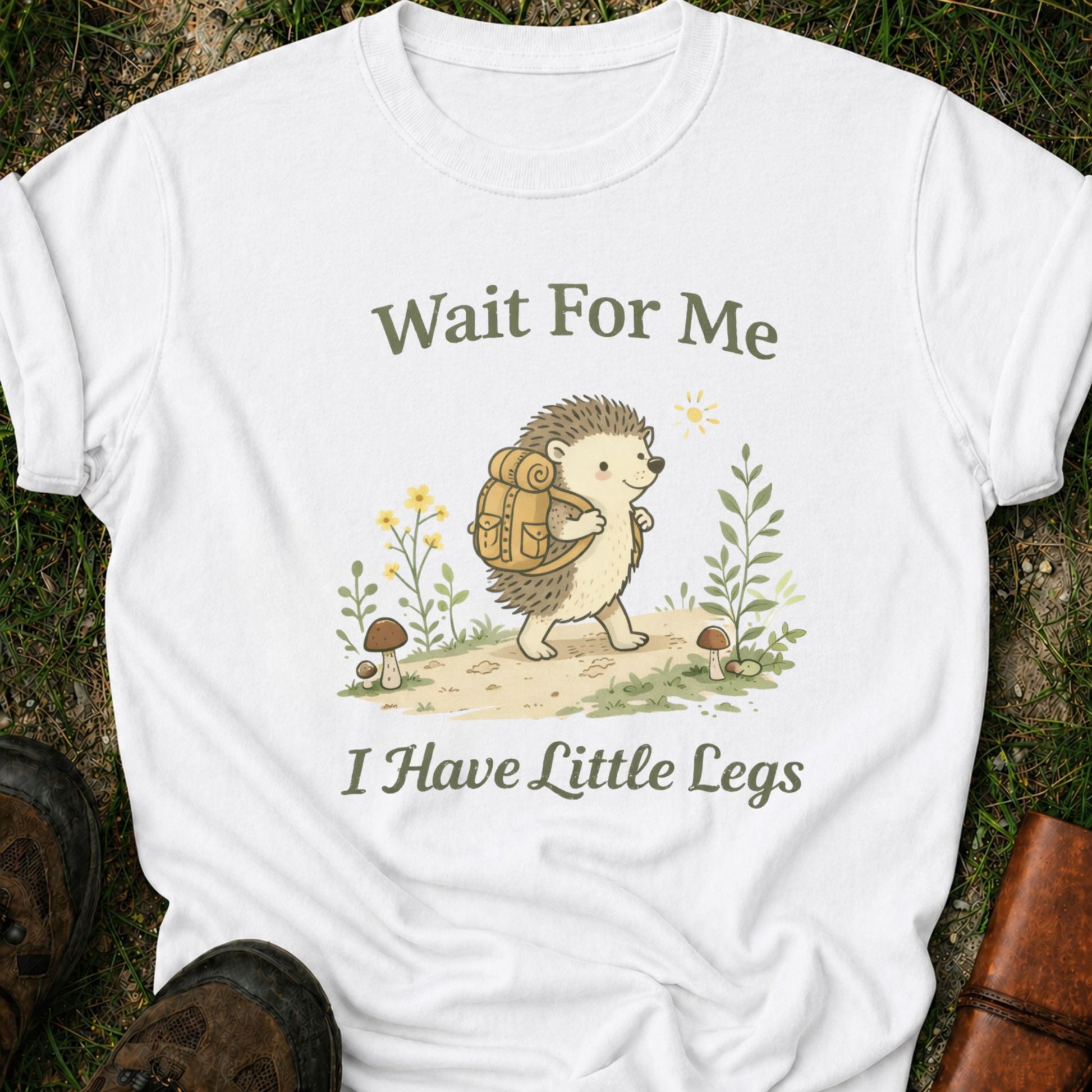 White t-shirt with a hedgehog with a hiking backpack strolls through a wildflower meadow, surrounded by mushrooms and greenery. Text reads "Wait For Me, I Have Little Legs."