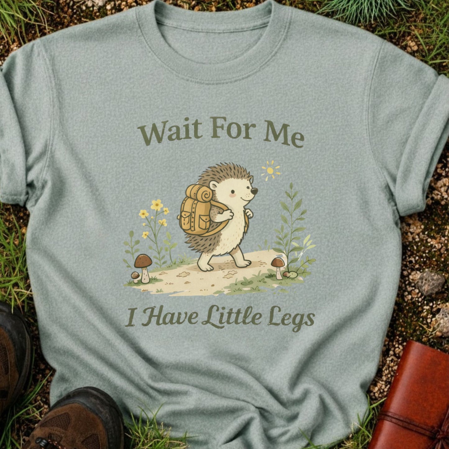 Green T-shirt with a hedgehog with a hiking backpack strolls through a wildflower meadow, surrounded by mushrooms and greenery. Text reads "Wait For Me, I Have Little Legs."