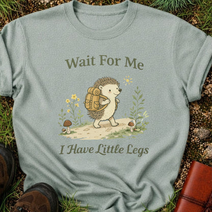 Green T-shirt with a hedgehog with a hiking backpack strolls through a wildflower meadow, surrounded by mushrooms and greenery. Text reads "Wait For Me, I Have Little Legs."
