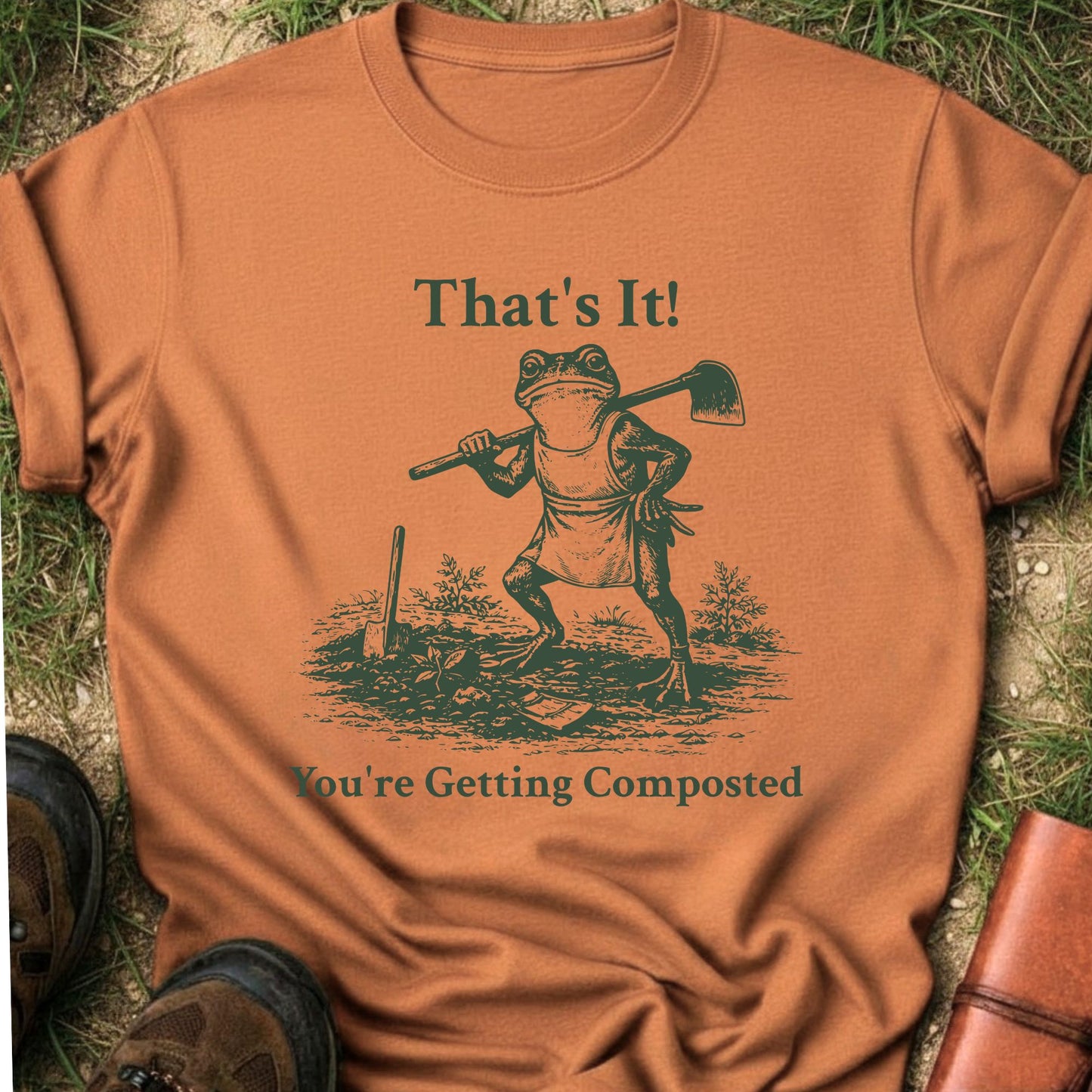 Orange Tshirt lying on a grassy surface.  Graphic is a frog with a hand on his hip holding a hoe with the tagline "That's It!, You're Getting Composted.