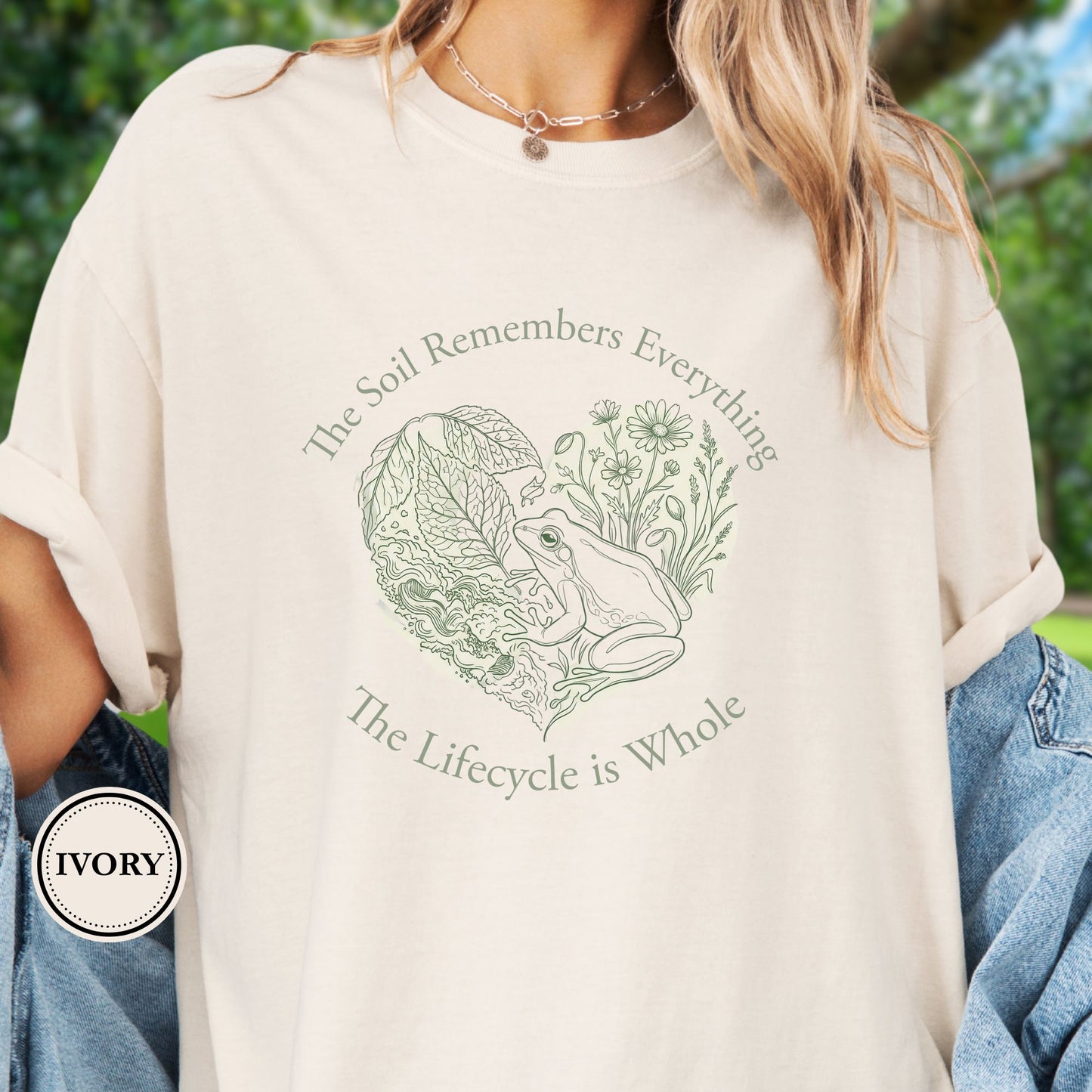 Model wears a Ivory Tshirt,  the graphic features a delicate line art of a frog, soil, leafy greens, and wildflowers with the phrase “The Soil Remembers Everything. The Lifecycle is Whole”
