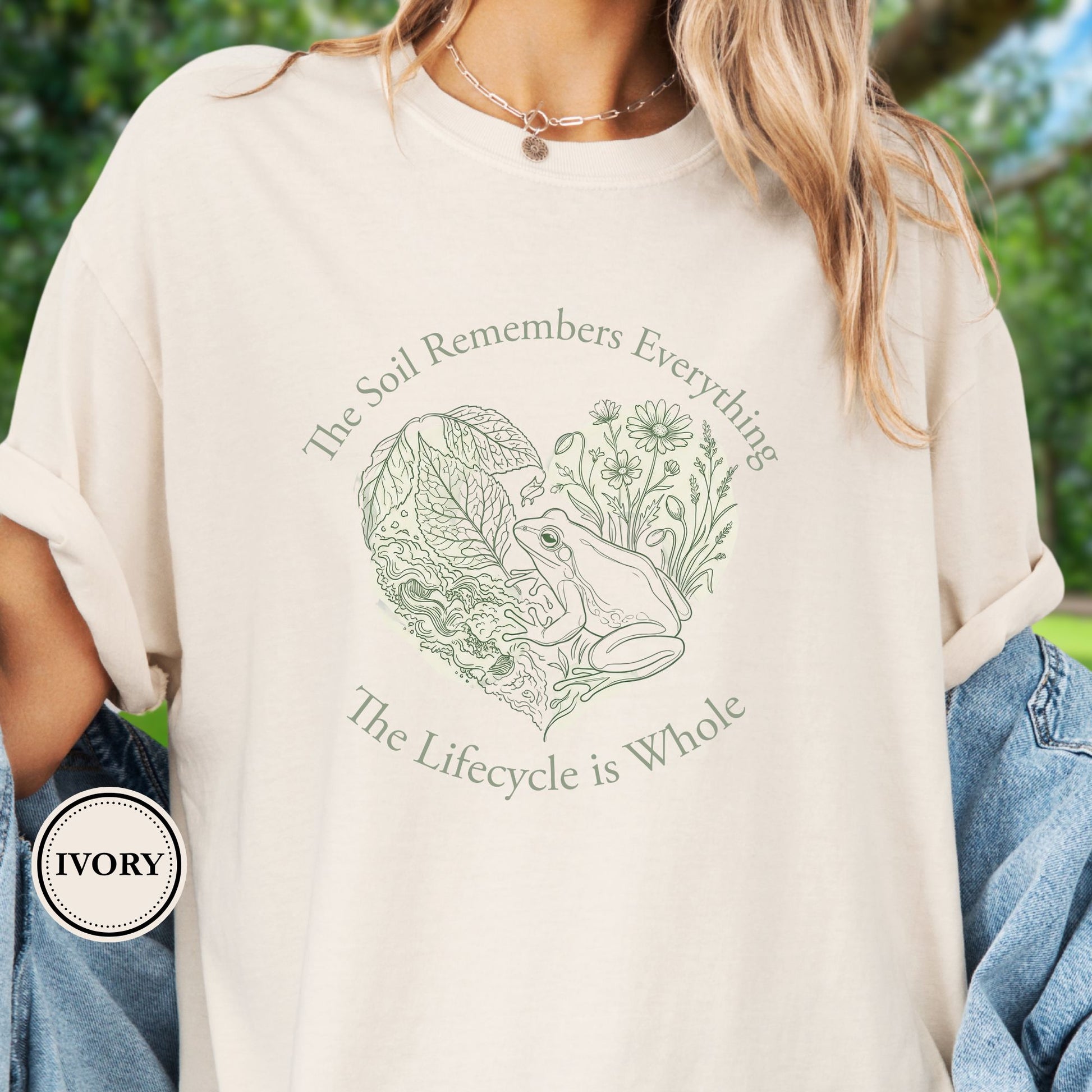 Model wears a Ivory Tshirt,  the graphic features a delicate line art of a frog, soil, leafy greens, and wildflowers with the phrase “The Soil Remembers Everything. The Lifecycle is Whole”