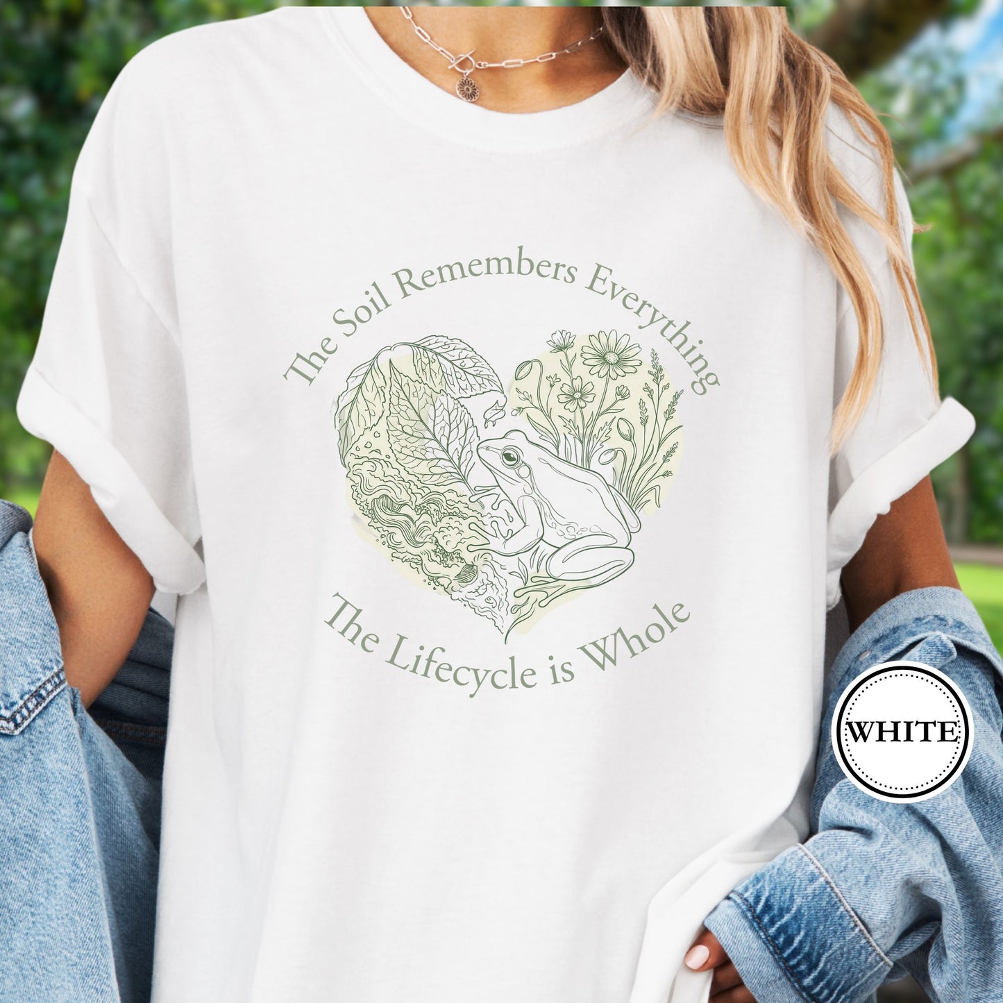 Model wears a White Tshirt,  the graphic features a delicate line art of a frog, soil, leafy greens, and wildflowers with the phrase “The Soil Remembers Everything. The Lifecycle is Whole”