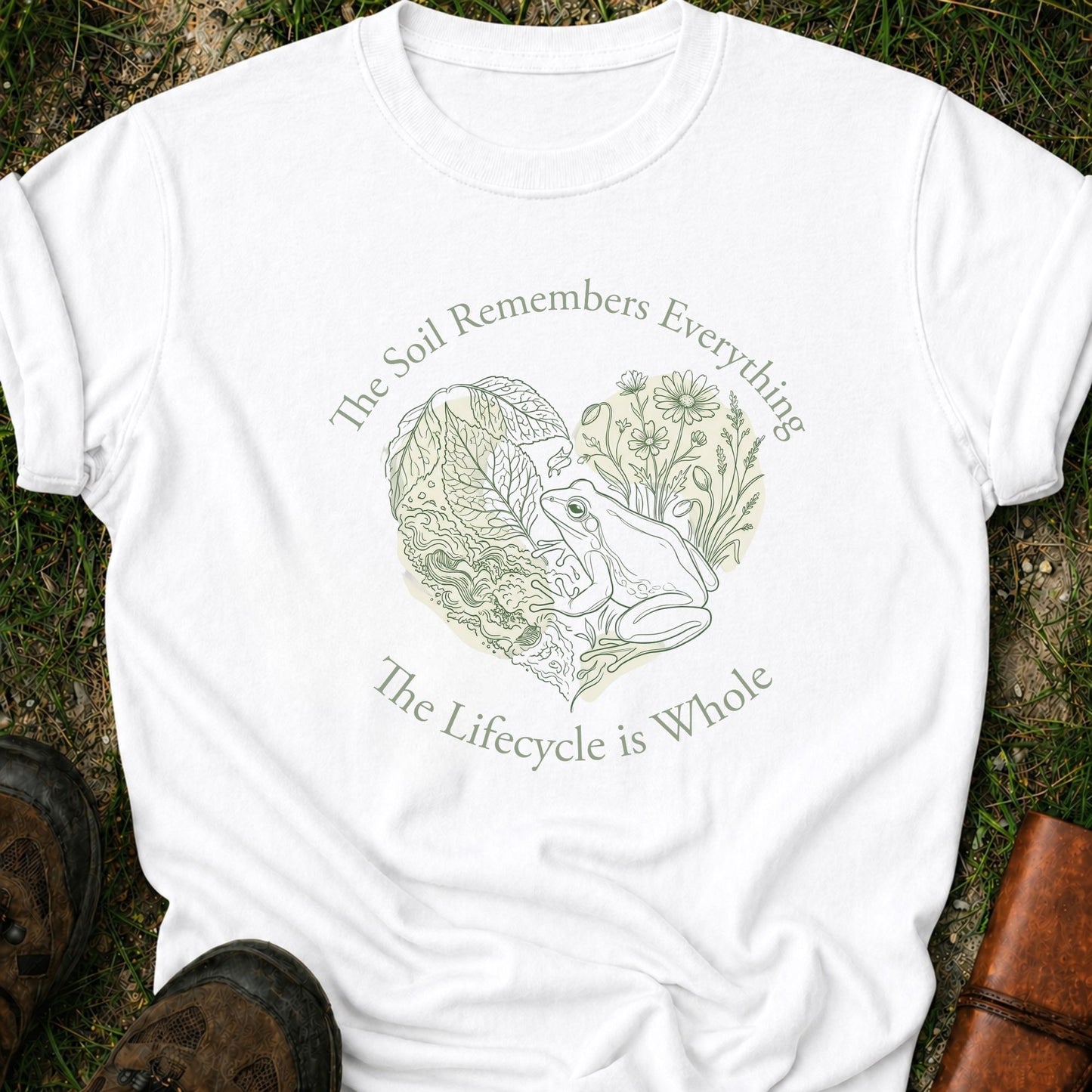 White Tshirt lying on a grassy surface,  the graphic features a delicate line art of a frog, soil, leafy greens, and wildflowers with the phrase “The Soil Remembers Everything. The Lifecycle is Whole”