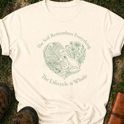 Ivory Tshirt lying on a grassy surface,  the graphic features a delicate line art of a frog, soil, leafy greens, and wildflowers with the phrase “The Soil Remembers Everything. The Lifecycle is Whole”