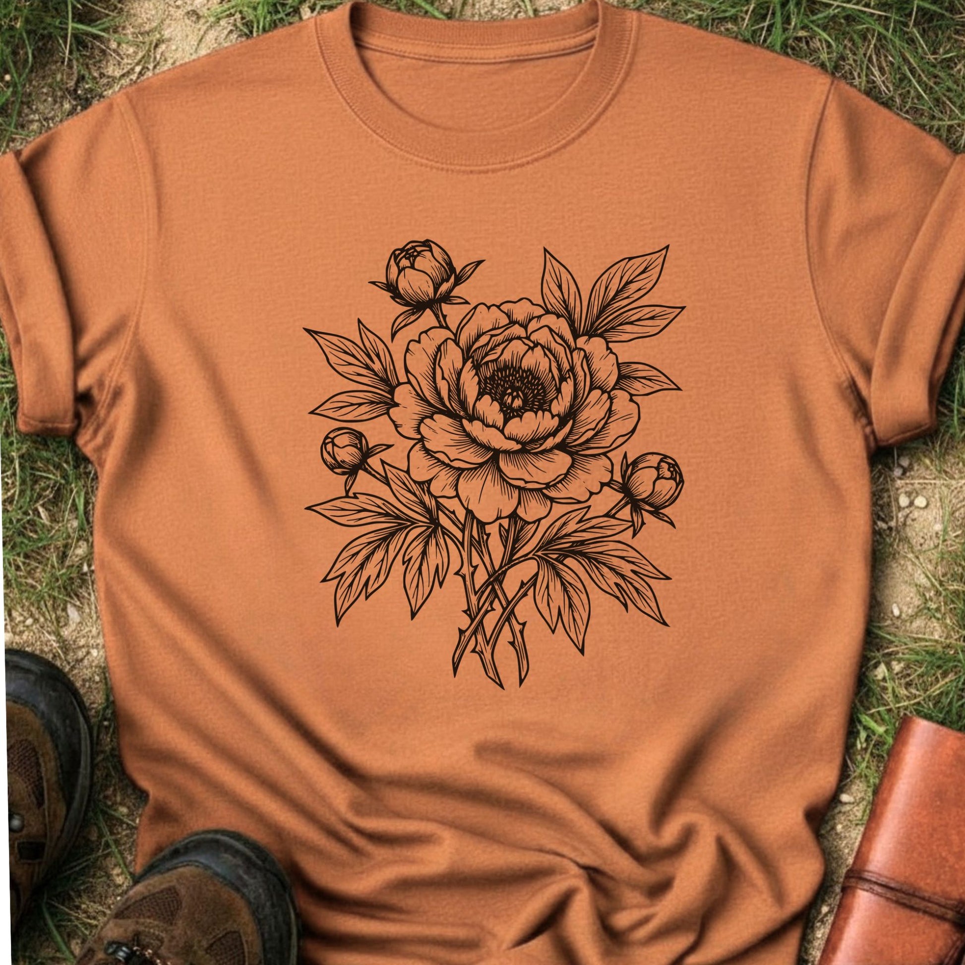 Orange Tshirt lying on a grassy surface. The black graphic on the shirt is an  botanical line art design featuring a detailed peony-style flower with leaves and buds.