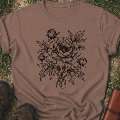 Brown Tshirt lying on a grassy surface. The black graphic on the shirt is an  botanical line art design featuring a detailed peony-style flower with leaves and buds.