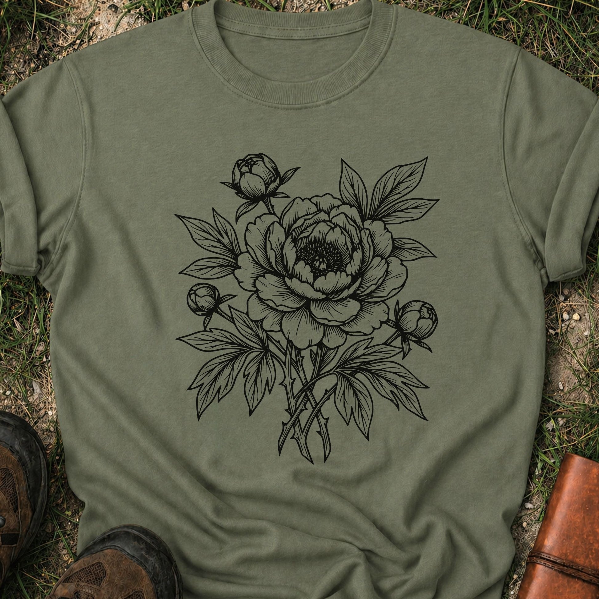 Green Tshirt lying on a grassy surface. The black graphic on the shirt is an  botanical line art design featuring a detailed peony-style flower with leaves and buds.
