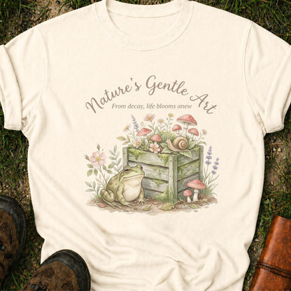 Ivory T-shirt lying on a grassy surface with a Whimsical cottagecore garden design featuring a frog, compost bin, mushrooms, wildflowers, and snail with the phrase “Nature’s Gentle Art. From decay, life blooms anew”