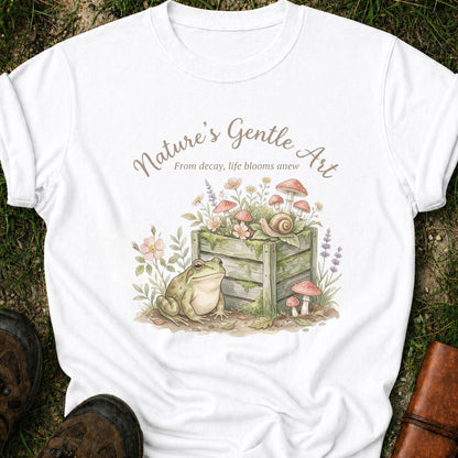 White T-shirt lying on a grassy surface with a Whimsical cottagecore garden design featuring a frog, compost bin, mushrooms, wildflowers, and snail with the phrase “Nature’s Gentle Art. From decay, life blooms anew”