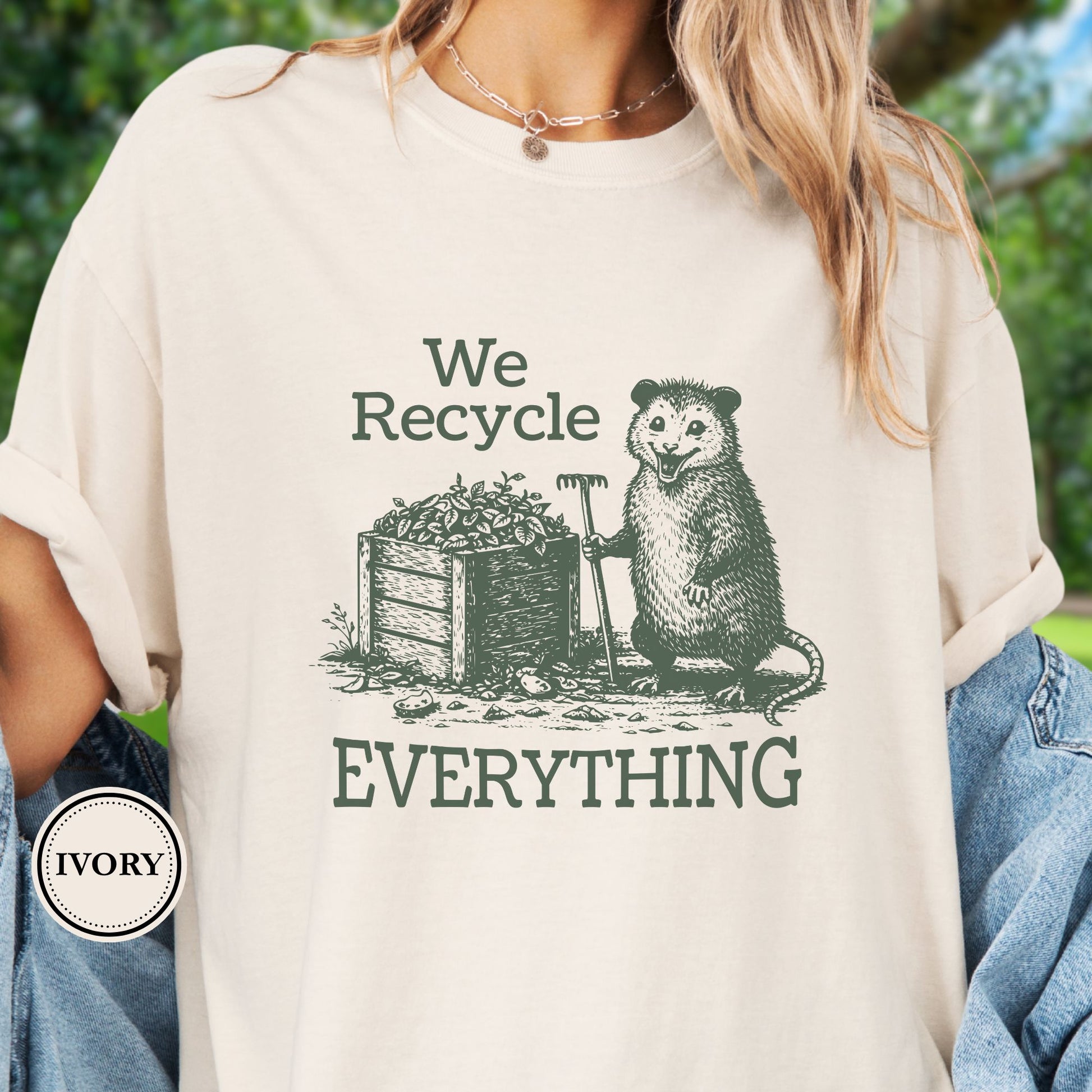 Model wears an Ivorty T-Shirt  featyrubg a vintage-style opossum beside a compost bin with the bold phrase “We Recycle Everything.” 