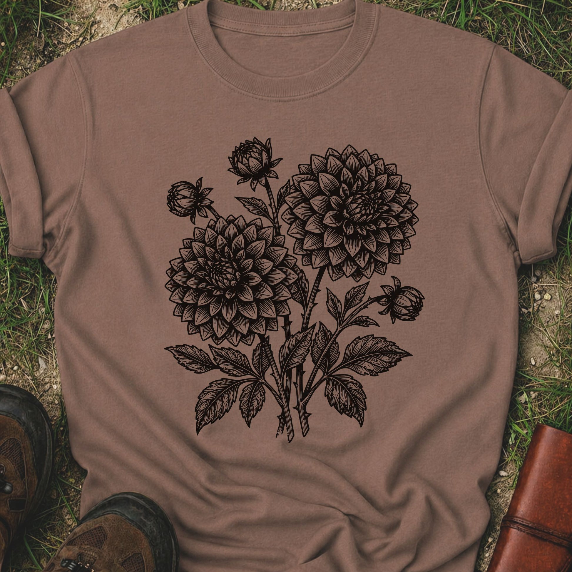 Brown T-Shirt laying on a grassy surface.  The graphic features a minimal black botanical line art design featuring dahlias with leafy stems and delicate floral details.