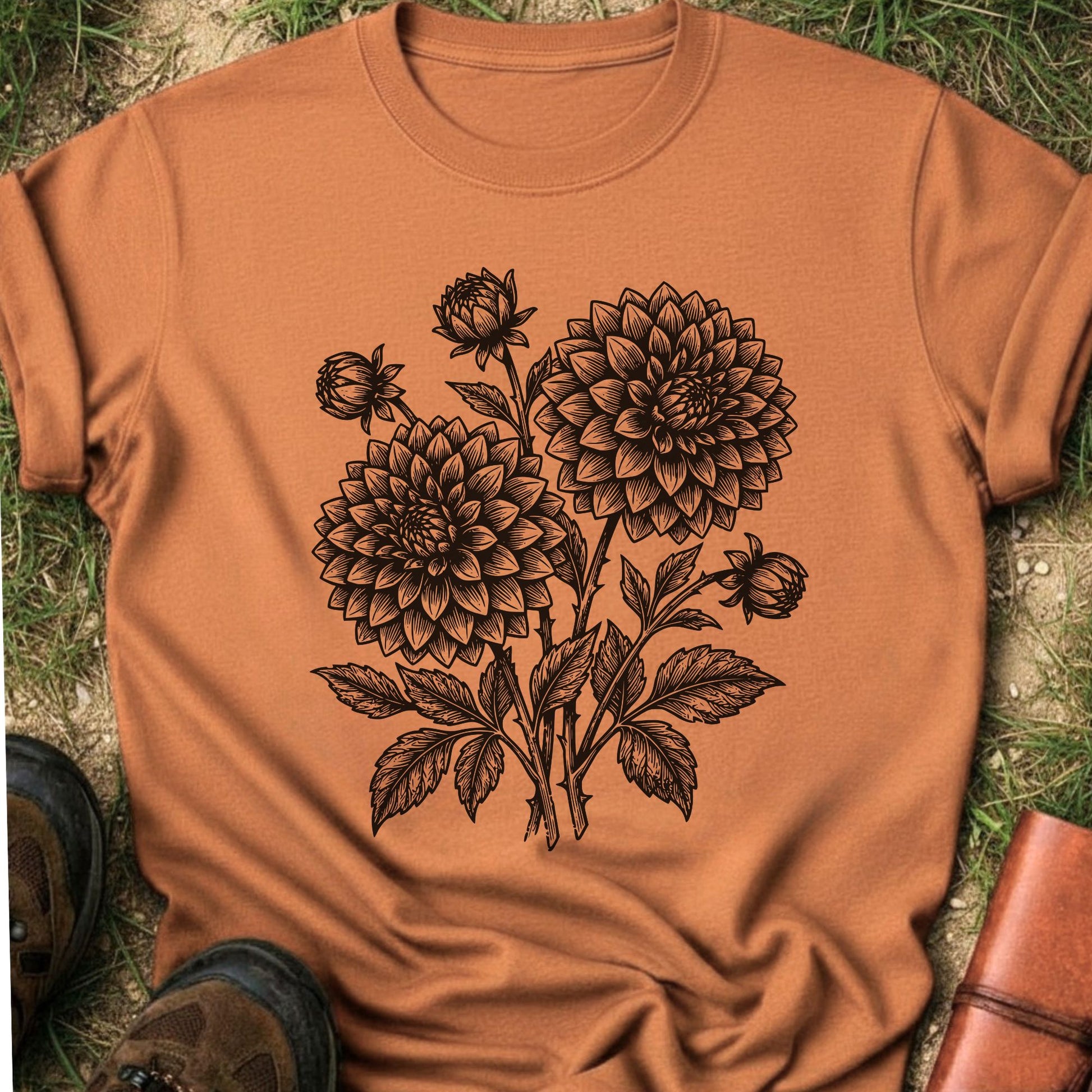 Orange T-Shirt laying on a grassy surface.  The graphic features a minimal black botanical line art design featuring dahlias with leafy stems and delicate floral details.