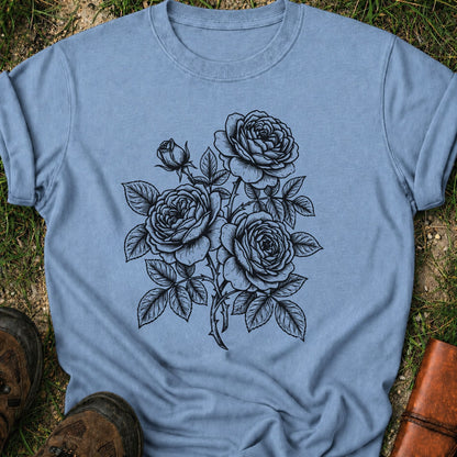 Blue Tshirt lying on a grassy surface.  Graphic shows minimal black botanical line art design featuring elegant roses with leafy stems