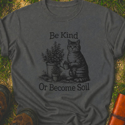 Dark Grey t-shirt lying on a grassy surface.  the graphic is a Funny vintage-style gardening cat design featuring a cat watering plants with the phrase “Be Kind Or Become Soil.”