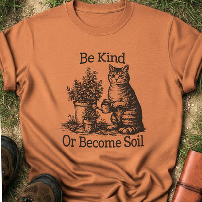  Orange t-shirt lying on a grassy surface.  the graphic is a Funny vintage-style gardening cat design featuring a cat watering plants with the phrase “Be Kind Or Become Soil.”